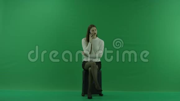 女孩看着左边坐在手提箱上在绿色屏幕上通过电话交谈视频的预览图