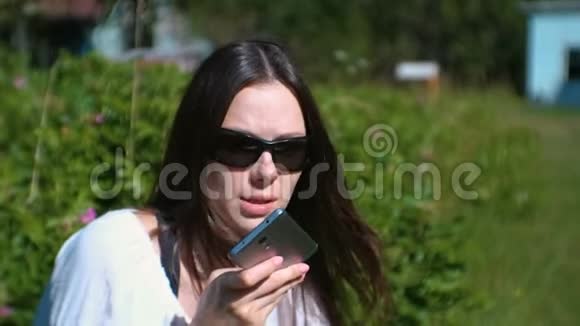 阳光明媚的一天美丽的黑发女人坐在公园里在手机上记录了一条语音信息视频的预览图