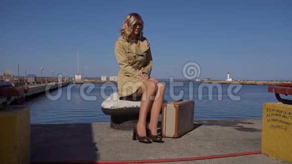 穿着老式西装的壕沟里迷人的年轻金发女人坐在游艇码头上视频的预览图