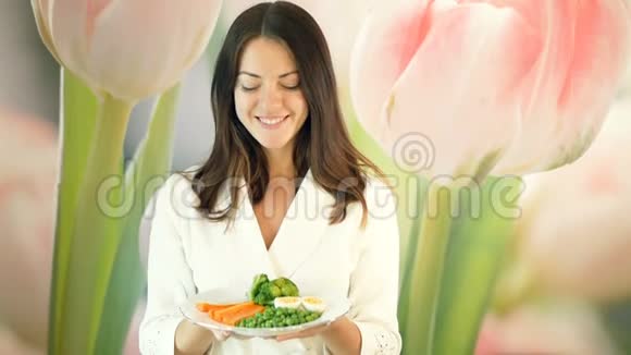 穿着白色长袍的漂亮女孩拿着一大盘煮熟的蔬菜向前推布鲁内特眨眼健康和健康视频的预览图