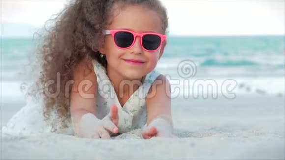 一个戴着粉红色眼镜的漂亮小女孩的特写肖像可爱的微笑着看着相机躺在沙滩上视频的预览图
