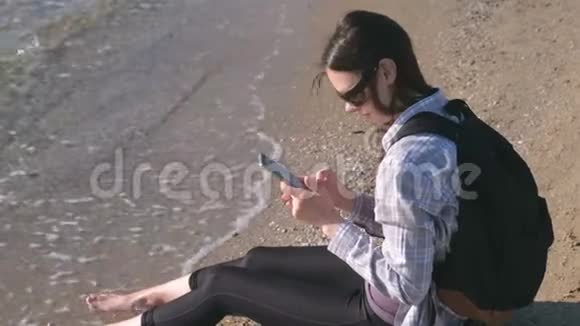 旅行者女孩带着背包坐在沙滩上在手机上输入一条信息视频的预览图
