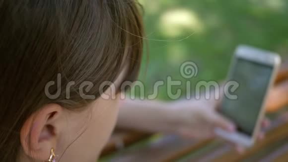 一个使用智能手机的年轻女孩正在公园的一家网上商店里的长凳上翻页年轻的千禧年视频的预览图