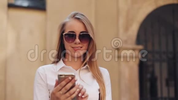 美丽的年轻商务女性穿着白色衬衫在市中心散步时使用智能手机夏季上网视频的预览图