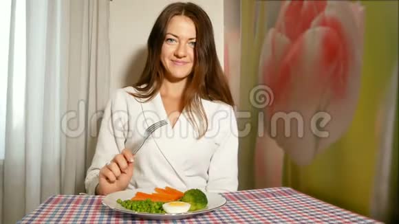 一个穿着白色长袍的漂亮女孩坐在厨房的一张桌子旁在叉子上捏了一块煮好的西兰花然后视频的预览图