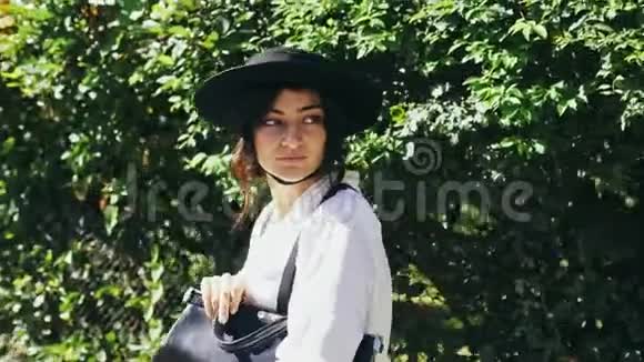在美丽的日子里一个戴着黑色帽子和吊带的女孩沿着绿色的灌木丛走着视频的预览图