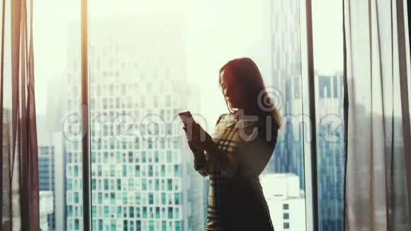 美丽的年轻女子站在窗边在美丽的夕阳和模糊的城市背景下使用智能手机视频的预览图