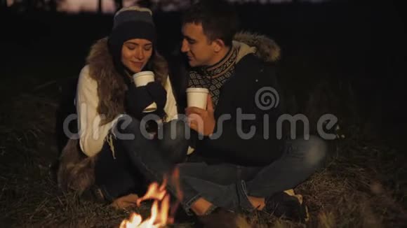 浪漫的情侣在篝火旁休息微笑爱抚喝茶视频的预览图