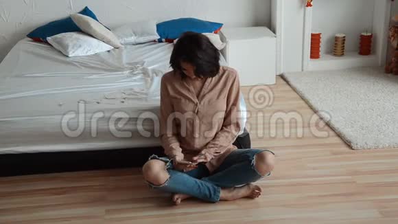 年轻的黑发女人坐在床边的地板上看着电话视频的预览图