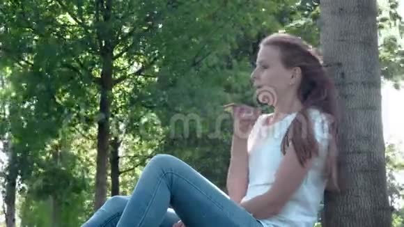 公园里穿着街服的年轻漂亮女孩手里拿着智能手机坐在树下的草地上视频的预览图