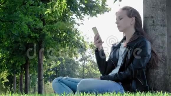 公园里穿着街服的年轻漂亮女孩手里拿着智能手机坐在树下的草地上视频的预览图