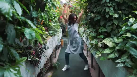 一个穿着围裙的漂亮女孩在温室里跳舞花店快乐又跳又笑视频的预览图