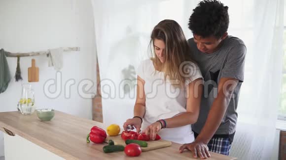 英俊的非洲男人正在拥抱白种人的女朋友同时帮助她早上在厨房准备食物视频的预览图