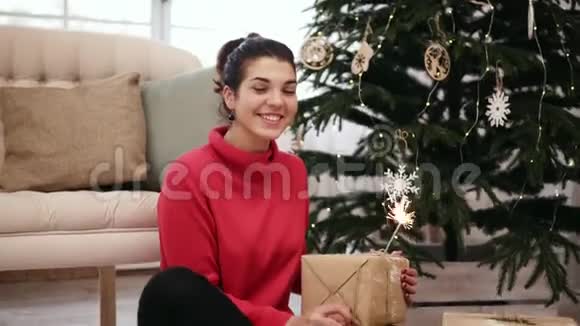 快乐迷人的年轻女子坐在圣诞树旁用火花庆祝圣诞节并向里面张望视频的预览图