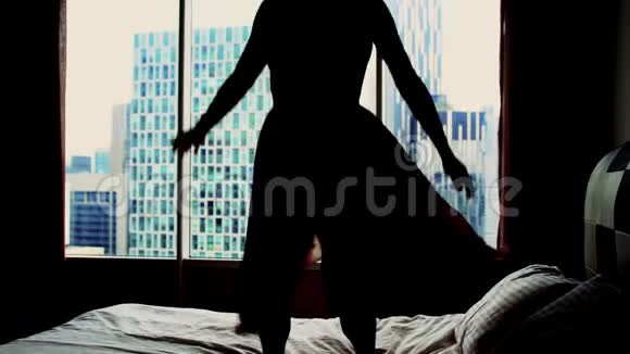 年轻的快乐女人的剪影穿着礼服在酒店床上跳着舞在背景建筑上的公寓里跳着视频的预览图