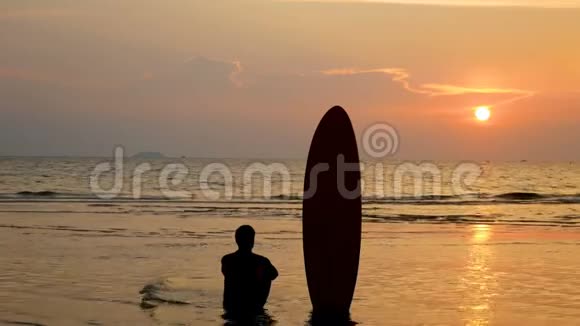 4K冲浪者坐在海边的剪影在热带海滩日落时有长长的冲浪板视频的预览图
