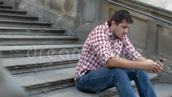 一个年轻悲伤的男人坐在老楼梯上在智能手机上使用应用程序视频的预览图