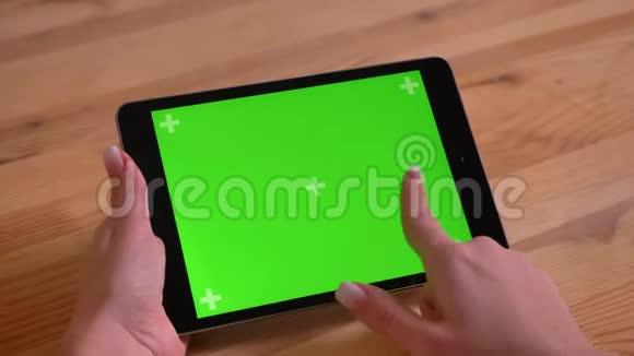 在木制桌面背景下通过绿色屏幕特写水平平板和女性手浏览视频的预览图