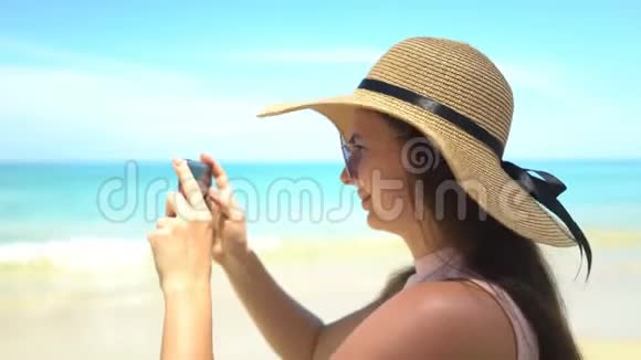 戴帽子的年轻旅行者女士在海边的智能手机拍照视频的预览图