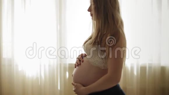 一个留着长发的怀孕女孩站在窗边抚摸着她的腹部慢动作视频的预览图