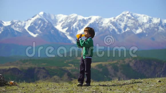 一个非常可爱的男孩在美丽的山脉背景上喝橙汁小男孩在山顶上放松视频的预览图
