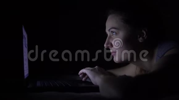 漂亮的年轻女人躺在床上在黑暗的房间里用笔记本电脑视频的预览图