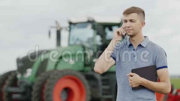 一位年轻成功的农民通过电话交流它站在一台大型拖拉机的背景下视频的预览图