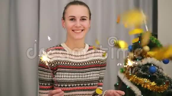 金色的纸屑落在年轻微笑的女人身上背景上有圣诞树和花环手里拿着火花视频的预览图