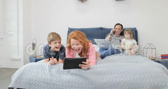 母亲和儿子一起用平板电脑观看躺在床上的滑稽视频视频的预览图
