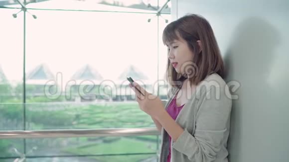 迷人的休闲快乐的年轻亚洲女性在国际机场使用和检查她的智能手机站在候机大厅视频的预览图