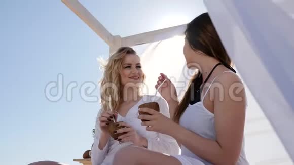 在背光阳光阳光下的快乐女孩女朋友从椰子壳喝热带鸡尾酒视频的预览图