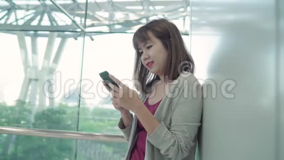 迷人的休闲快乐的年轻亚洲女性在国际机场使用和检查她的智能手机站在候机大厅视频的预览图