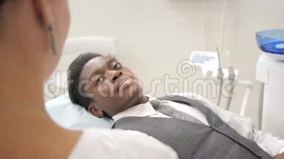女牙医检查并咨询病人年轻的非裔美国男性病人在牙科诊所的椅子上医药视频的预览图