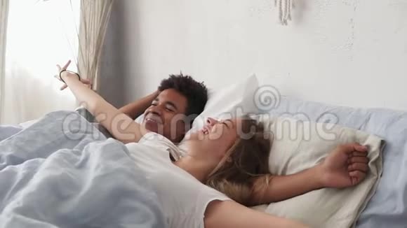 快乐的多种族夫妇躺在床上大笑早上一起醒来有魅力的年轻男女视频的预览图