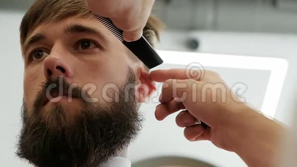 一位时髦的大胡子大胆男子用电动剃须刀修剪庙宇的特写理发师视频的预览图
