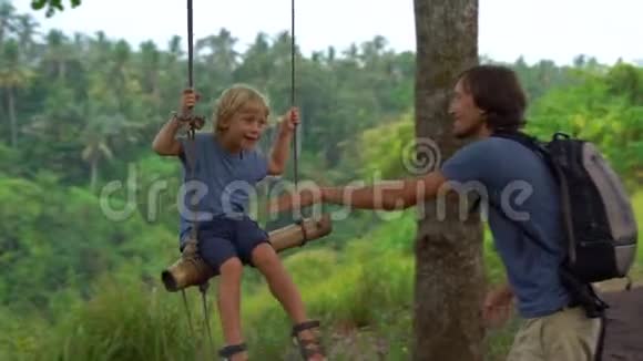 一个年轻人在热带公园的秋千上摆动他的小儿子的慢镜头儿童概念视频的预览图