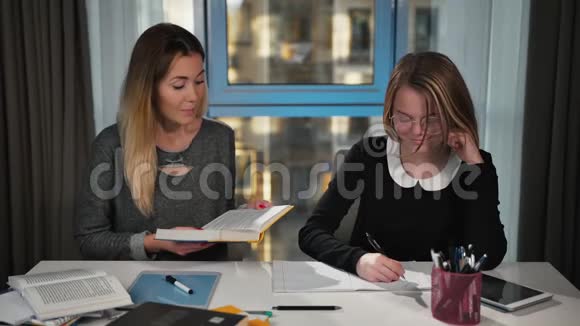 快乐的老师或妈妈教女学生读书女学生在练习本上写字微笑家庭视频的预览图