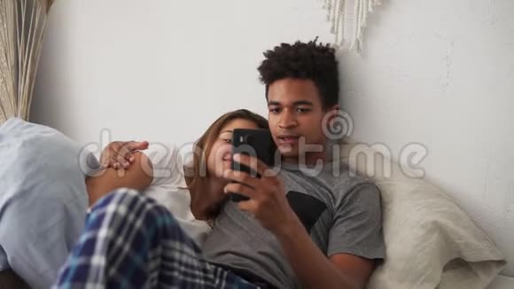 快乐的多民族年轻夫妇在卧室里放松智能手机互相拥抱慢镜头视频的预览图