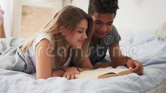 幸福微笑的多民族夫妇躺在床上一起在家里卧室看书慢镜头视频的预览图