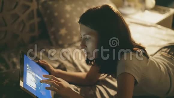 躺在床上的年轻微笑女子晚上在家里用数码平板电脑观看旅行照片视频的预览图