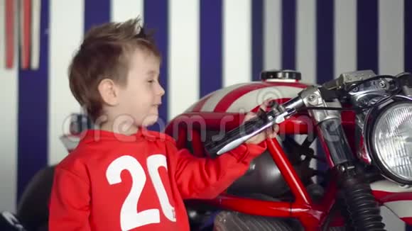 小可爱的男孩坐在红色的大摩托车上这个小男孩在演播室里以美国风格微笑关闭视频的预览图