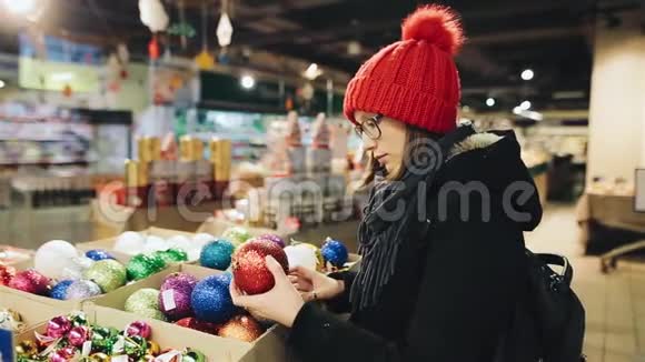 年轻漂亮的白种人女孩戴着眼镜和一顶红帽子在市场商店里挑选新年树的球视频的预览图