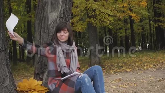 从笔记本上取一片叶子在公园里的草地上坐着一位迷人的年轻快乐的微笑女人在笔记本上记笔记视频的预览图