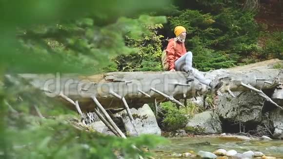 戴着帽子背着背包坐在圆木上的漂亮女游客坐在针叶林的一条山河上视频的预览图