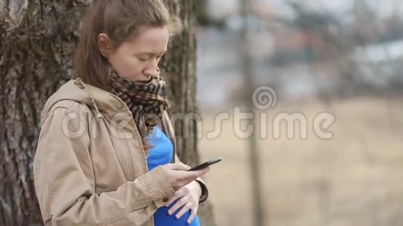 年轻的孕妇穿着浅棕色的外套和蓝色的t恤深情地用手轻轻地滚动着智能手机视频的预览图