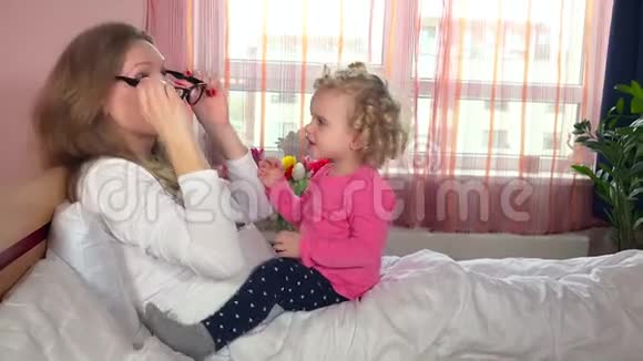 可爱的女儿试着用妈妈的眼镜坐在床上幸福的家庭女孩视频的预览图