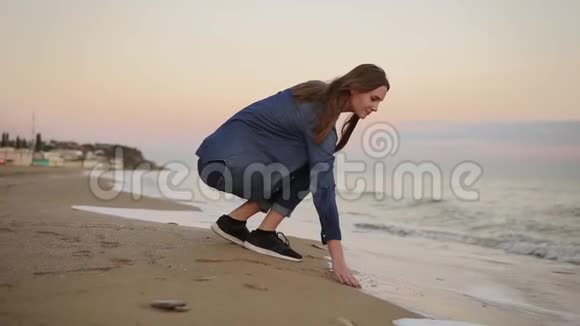 美丽的年轻女子坐在海边在日落时用手在沙滩上尝试水温视频的预览图