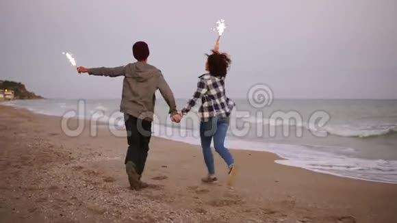 在日落时分年轻的多民族夫妇手持燃烧的闪闪发光的蜡烛在海边奔跑慢动作视频的预览图
