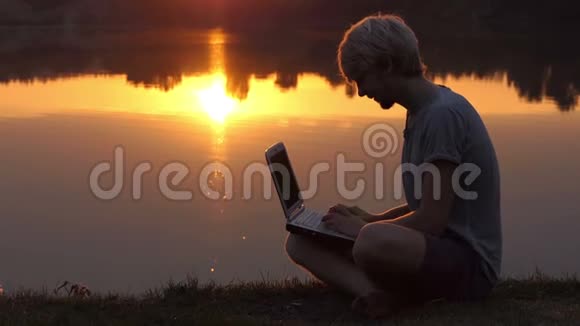 在日落时分在河岸上的笔记本电脑上印有创意的人视频的预览图