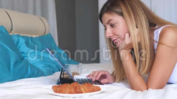 年轻女子手提电脑在家里的床上吃牛角面包喝咖啡视频的预览图